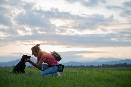 Happy cheerful young woman kneeling down to cuddle her cute black labrador puppy as they are outside on a walk in green meadow.の写真素材