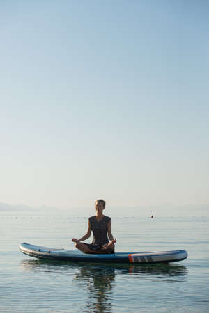 Young woman sitting on a paddle board floating on a calm morning sea, meditating in a lotus position.の写真素材