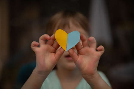Little toddler child holding a heart shaped paper in the colors of ukrainian flag - yellow and blue - in a conceptual image of a problem and horror of ukrainian war.の写真素材