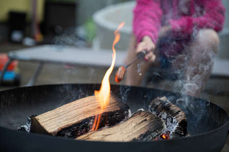 Closeup view of a woman grilling a hotdog in a stick above an open fire in an iron fireplace outside...