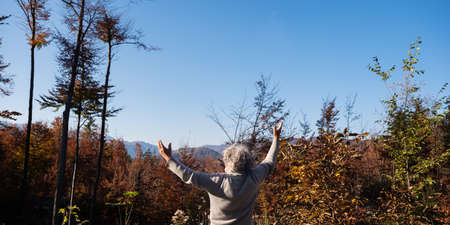 Senior man with grey curly hair celebrating the fall of his life with arms raised high standing in...