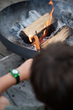 Over the head view of a boy grilling hot dog on a stick above an open fire in a cast iron grill in domestic backyard.の写真素材