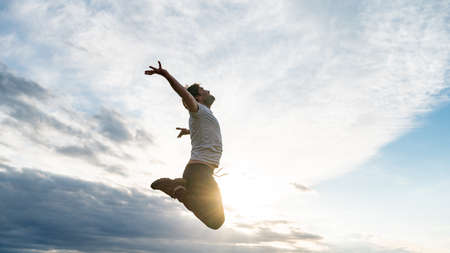Excited young man in casual clothes jumping high up in the air, oppening his arms towards the sky backlit by the evening sun.の写真素材