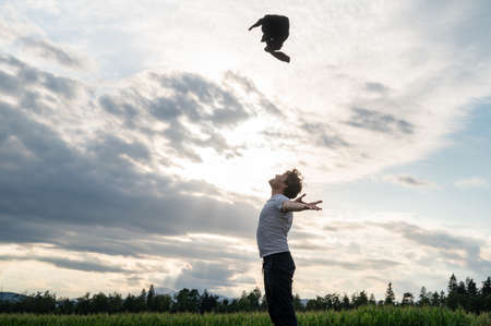 Cheerful young man standing in nature under beautiful evening sky throwing his sweater high up in the air in excitement.の写真素材