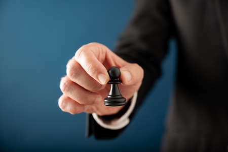Closeup view of hand of a businessman holding black chess pawn piece over blue background with copy space in a conceptual image of employment and leadership.の写真素材