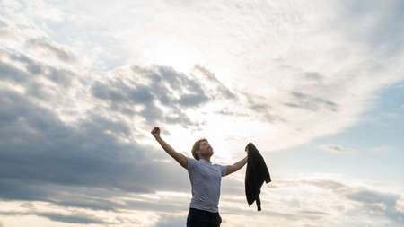 Proud young man standing under cloudy evening sky celebrating his life and success with open arms with clenched fists.の写真素材