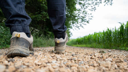 Low angle closeup view of male legs in hiking shoes walking on a country road running through green fields and nature.の写真素材