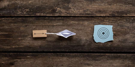 Origami boat pulling wooden peg with arrow drawn on it towards a blue paper with target sign. Conceptual image of vision and clear goals. Over rustic wooden background.の写真素材