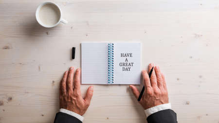 Top view of male hands in business suit placed next to an open notepad with Have a great day sign written in it. Over white wooden background with cup of coffee.の写真素材