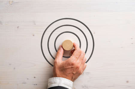 Hand of a businessman placing blank wooden cut circle in the middle of a target drawn on white...