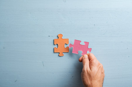 Male hand matching two blank puzzle pieces in orange and pink pastel colours. Over blue wooden...