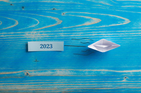 Origami made paper boat pulling a 2023 sign cut into a wooden plate. Over textured blue background.の写真素材