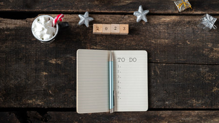 Top view of 2023 sin on wooden blocks next to a To do list in a notepad and holiday hot cocoa drink placed on wooden desk decorated with christmas ornaments.の写真素材