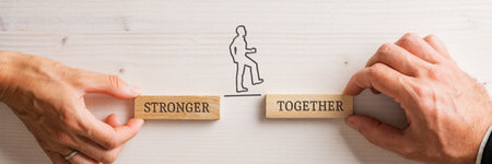 Hands of a businessman and businesswoman holding two wooden pegs with a Stronger together sign for a silhouetted businessman to walk confidently between the two. Wide view image.の写真素材