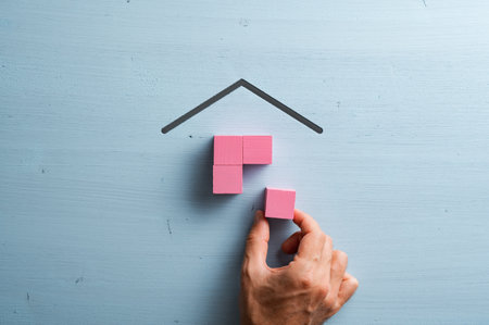 Male hand assembling a house made of pink wooden blocks over blue background in a conceptual image of home ownership and mortgage.の写真素材