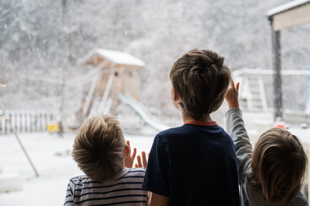 View from behind of three children, siblings, looking out the window into a beautiful winter nature with snow falling down the sky.の写真素材