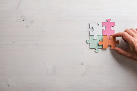 Male hand joining four colorful puzzle pieces over wooden background with plenty of copy space on the left side of an image.の写真素材