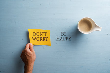 Dont worry Be happy sign written partially on yellow piece of paper and partiall cut into w pastel blue wooden background. Cup of coffee placed next to the text. Top view.の写真素材