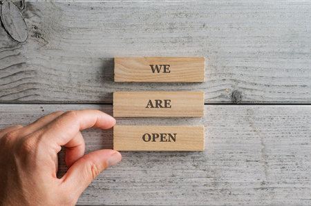 Male hand assembling a We are open sign written on three stacked wooden blocks placed over white wooden background.の写真素材
