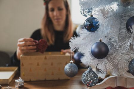 Young woman packing christmas present by the white holiday tree with shiny blue baubles. Focus to the balls on the tree.の写真素材