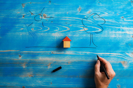 Male hand drawing a tree and sun around a house made of wooden blocks over textured blue background in a conceptual image of home and home ownership.の写真素材