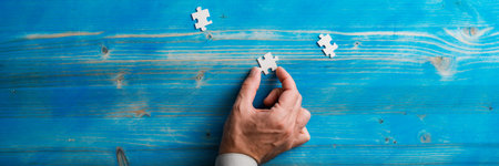 Male hand holding a blank puzzle piece over blue wooden background with two other pieces lying around. Wide view image.の写真素材