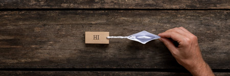 Wide view image of male hand holding an origami made paper boat pulling a wooden peg with Hi sign on it. Over rustic wooden background.の写真素材