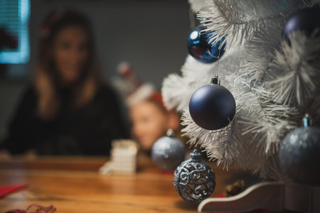 Mother and daughter making holiday decoration by a white christmas tree decorated with shiny blue balls. Focus to the bauble decor.の写真素材
