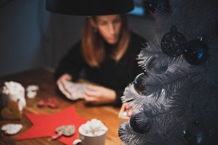 Closeup view of white christmas tree decorated with shiny blue holiday baubles placed on domestic table with blurred young woman packing holiday gifts in background.の写真素材