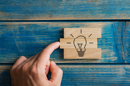 Male finger pushing a wooden peg into a stack of them to complete a light bulb shape drawn on them in a conceptual image of idea, vision and business startup. Over blue wooden background.の写真素材