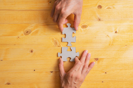 Male and female hand joining two matching puzzle pieces over yellow wooden background in a conceptual image of merger, teamwork and cooperation.の写真素材