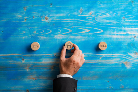 Hand of a businessman or real estate agent placing a wooden cut circle with a house icon on it in the middle between Sell and Buy signs. Over textured blue wooden background.の写真素材