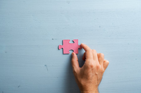 Male hand placing a blank pink puzzle piece over a plain pastel blue wooden background.の写真素材