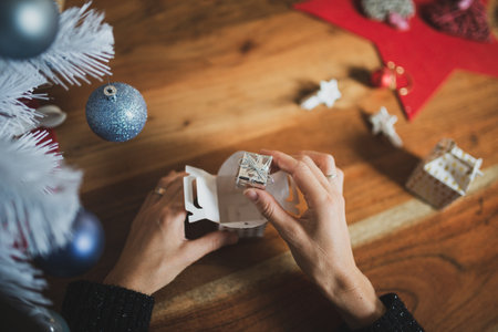 Top view of female hands packing a small giftbox present into a bigger box next to a white christmas tree with blue holiday baubles.の写真素材