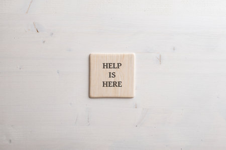 Wooden tile with a Help is here sign written on it placed over plain simple wooden background.の写真素材