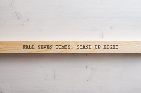 Motivational Fall seven times, stand up eight sign written on wooden slat placed horizontally over plain simple wooden background.の写真素材