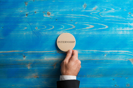 Male hand in business suit placing a wooden cut circle with an Internship word on it over a blue wooden background.の写真素材