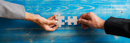 Wide view image of businessman and businesswoman, partners, matching and joining two blank puzzle pieces in a conceptual image. Over textured blue wooden background.の写真素材