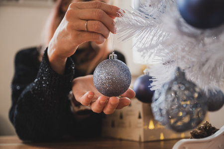Closeup view of female hands about to hang a shiny blue holiday bauble on white christmas tree in a domestic life image.の写真素材
