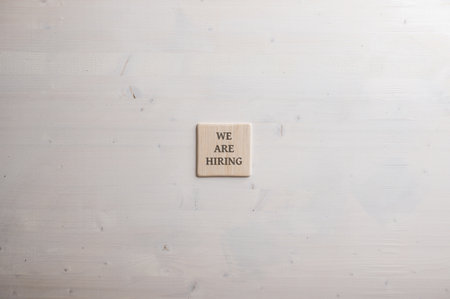 Wooden tile with a We are hiring sign on it placed over plain simple wooden background.の写真素材
