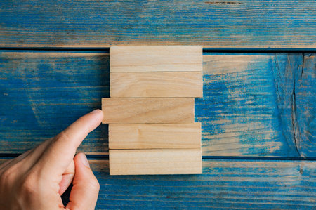Male finger pushing a simple blank wooden peg into a stack of them in a conceptual image of building a business, startup and stability. Over textured blue wooden background.の写真素材