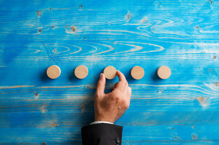 Hand of a businessman placing five simple blank wooden cut circles over a textured blue wooden background.の写真素材