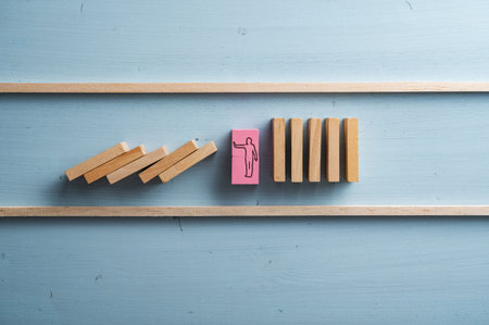 Pink wooden domino with a silhouette of a man making a stop gesture placed in the middle of collapsing dominos to interrupt and prevent crisis.の写真素材