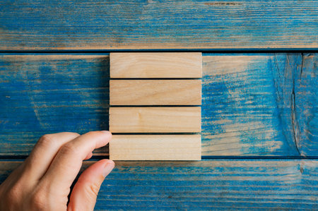 Male hand stacking four blank wooden pegs over a textured blue wooden background.の写真素材