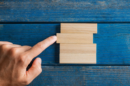 Conceptual image of business startup and stability - male finger pushing simple blank peg into a stack of them. Over textured blue wooden background.の写真素材