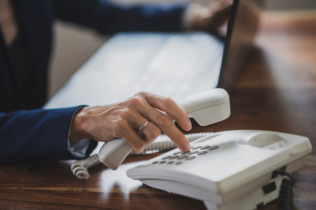 Businesswoman working on laptop computer dialing a telephone number using white landline phone on her desk.の写真素材