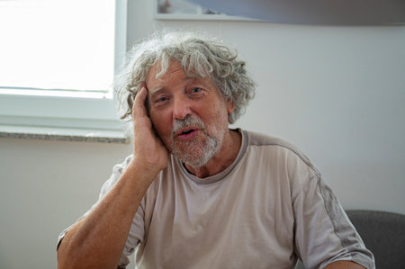 Authentic senior man with gray curly hair leaning his head on his arm with his mouth as if he is speaking or singing.の写真素材