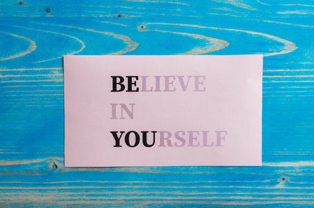Believe in yourself sign fading into a Be you message written on pink paper placed over textured blue wooden background.の写真素材
