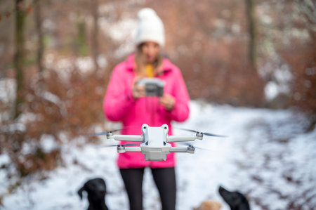 Young woman in pink winter jacket standing in snowy forest using a controller to direct and manage a drone floating in front of her. Focus to the drone.の写真素材