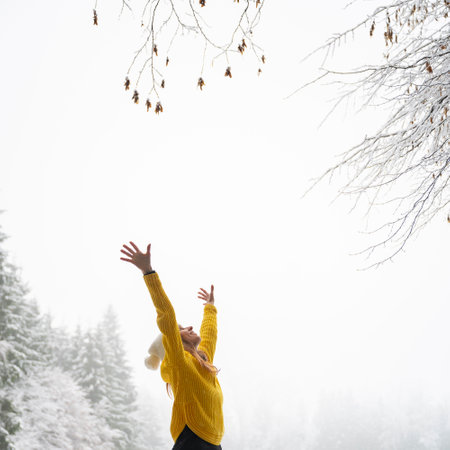 Profile view of a cheerful young woman in bright yellow sweater enjoying and celebrating life with her arms raised high in the air while standing outside in snowy winter nature.の写真素材